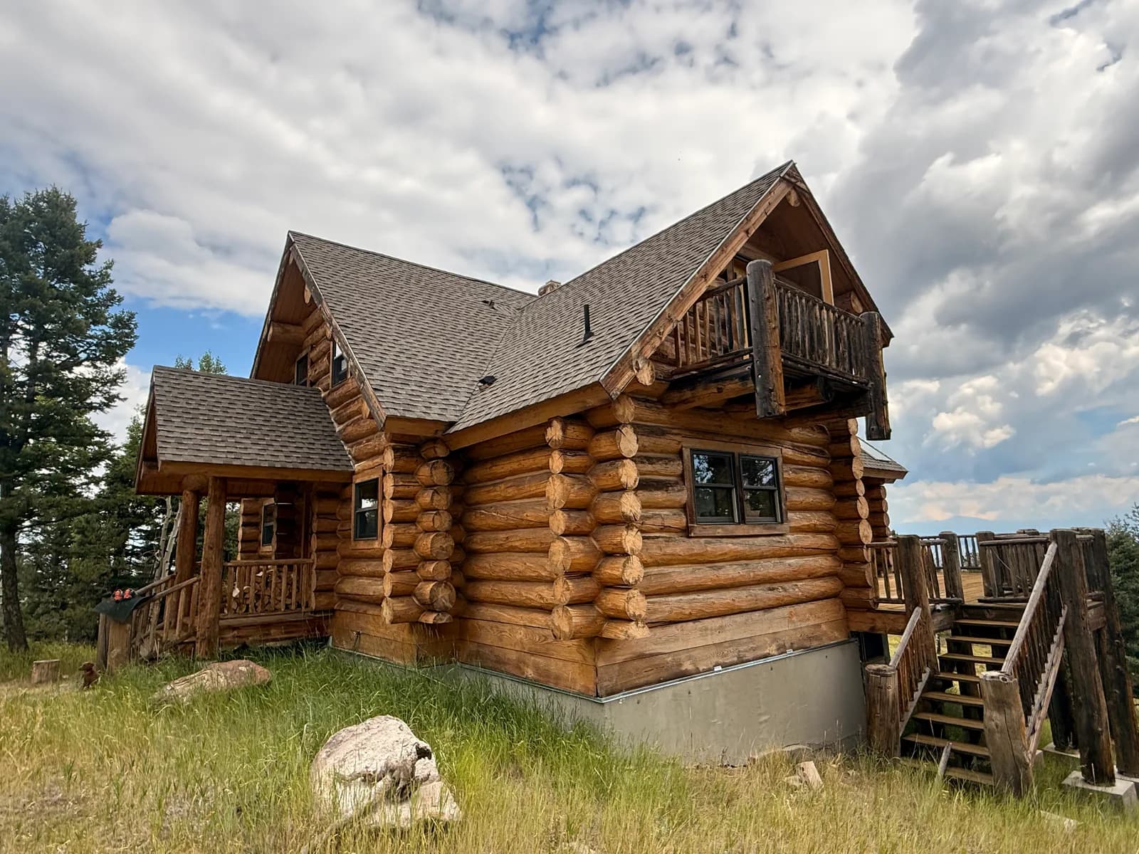 Large-diameter full-scribe log home with a freshly applied honey stain, gable rooflines, and a wraparound deck under a blue Colorado mountain sky