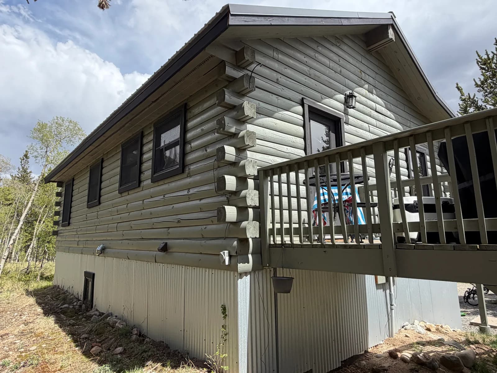 Sage-green stained log cabin with black window trim, a wood deck, and protective metal skirting in a forested Colorado mountain setting