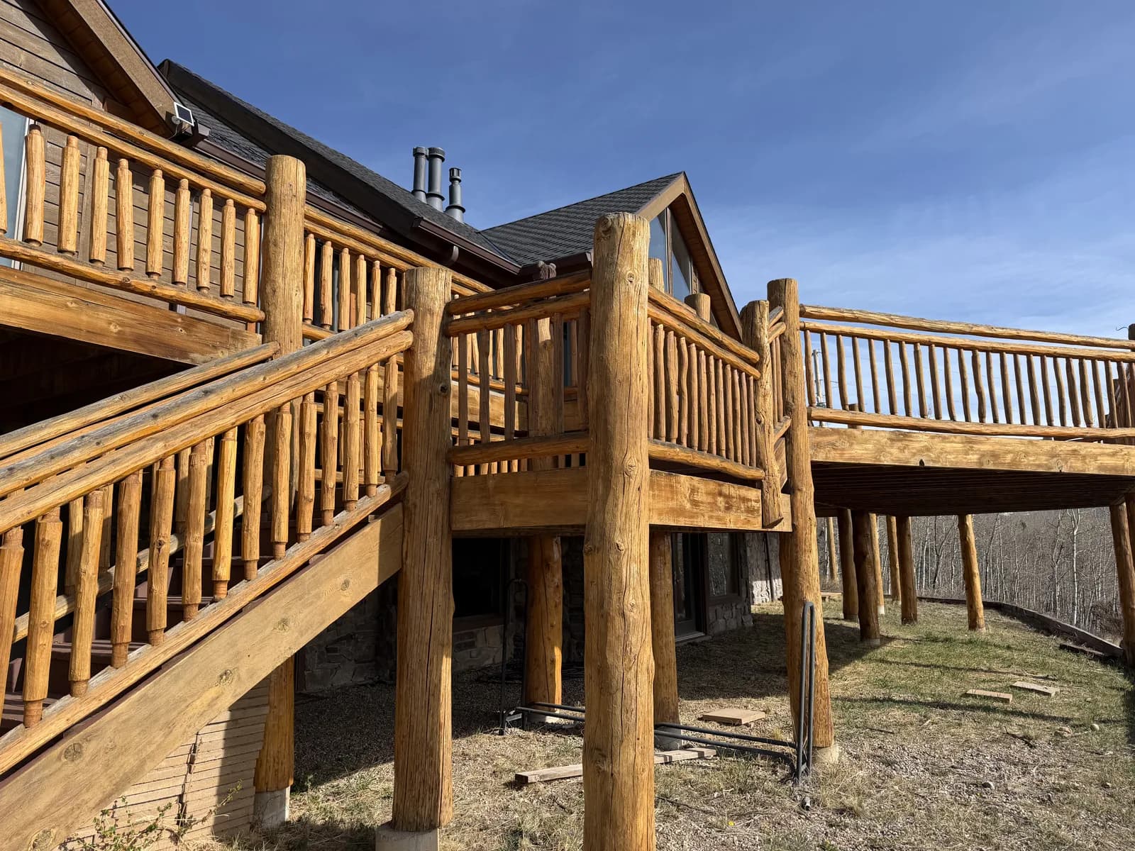 Multi-Level Log Deck & Railing Restoration Large multi-level log home deck with log railings and stairs, freshly stained to a warm golden tone, on a forested Colorado hillside