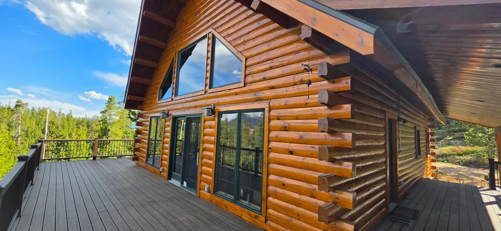 Log Home Staining & Deck Finishing Freshly stained honey-toned log home with green window trim and a large wraparound deck against a forested Colorado mountain backdrop