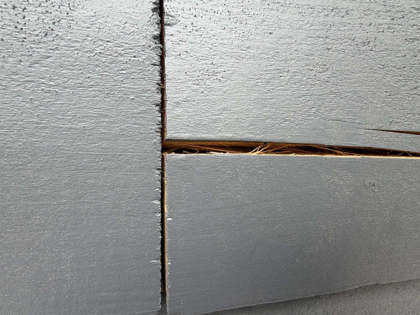 Open uncaulked vertical butt joint between two grey log siding boards with the horizontal reveal below packed with pine needles and debris