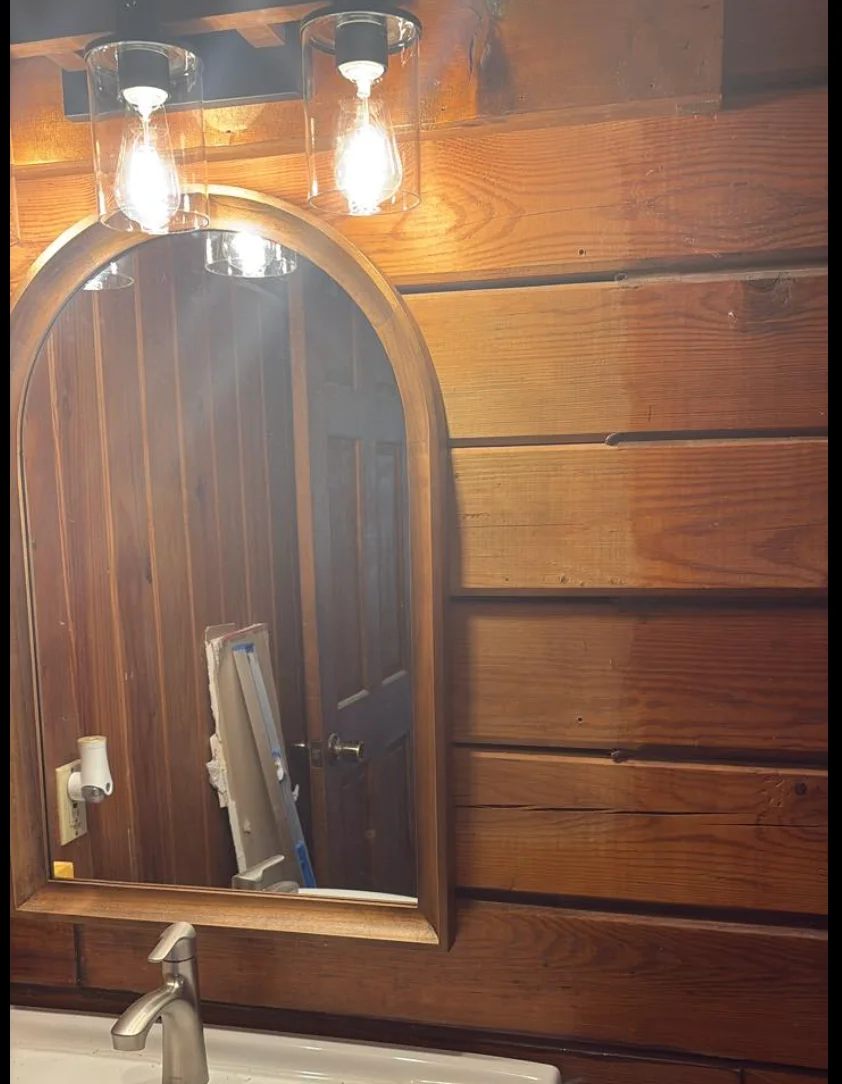 Log home bathroom interior showing water staining on D-log walls near the vanity area, with visible discoloration on the flat-milled log surfaces behind a mirror and light fixture